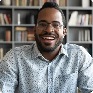 Black man with beard and glasses wearing a patterned button down shirt smiling in front of a book case