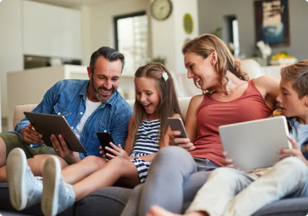 A mom, dad, son, and daughter sitting on a couch looking at their electronic devices and laughing