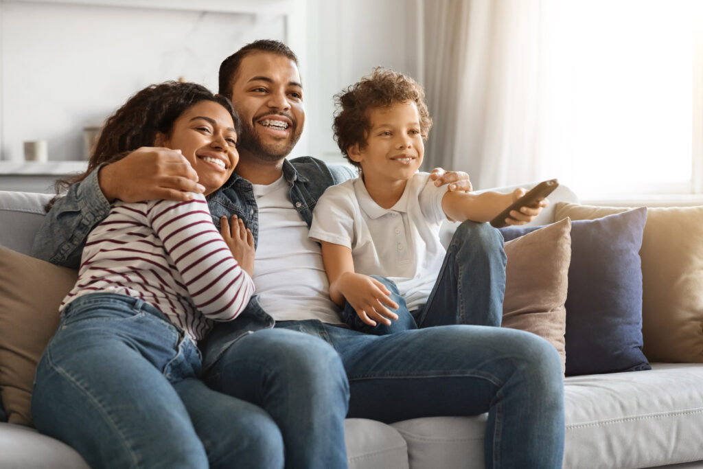 African american family with their preteen son cuddling and watching free cable tv together at home, loving parents and male kid relaxing on couch in living room, boy holding remote controller