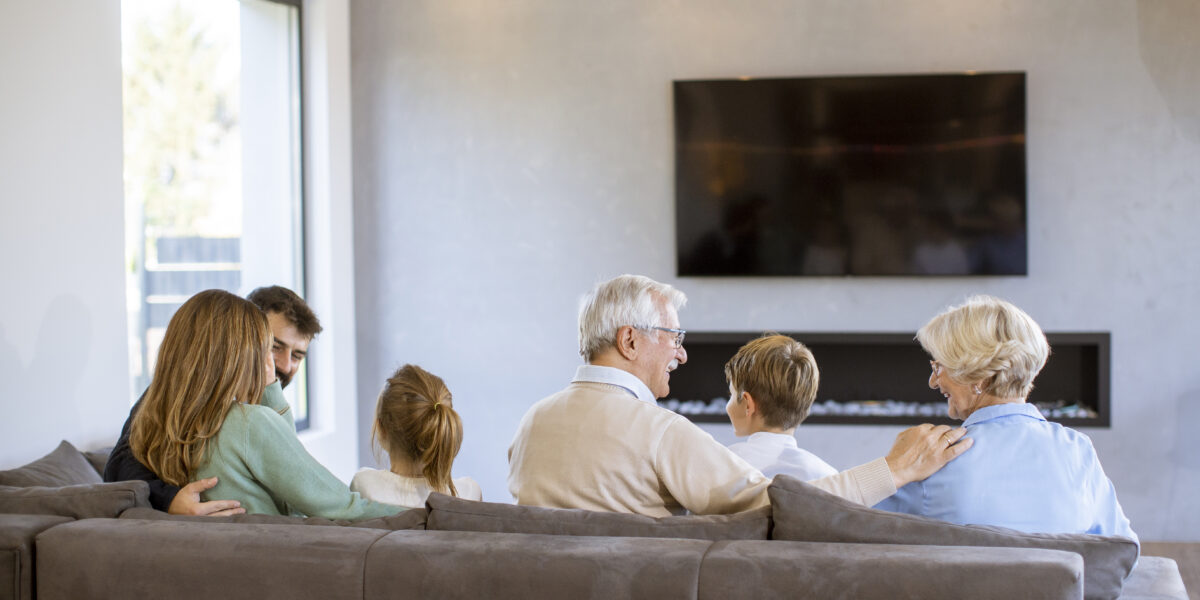 A multi-generational family sitting together on a sofa in a modern living room, enjoying quality time while watching free cable TV. The group includes grandparents, parents, and children, all facing the television, creating a warm and cozy atmosphere.