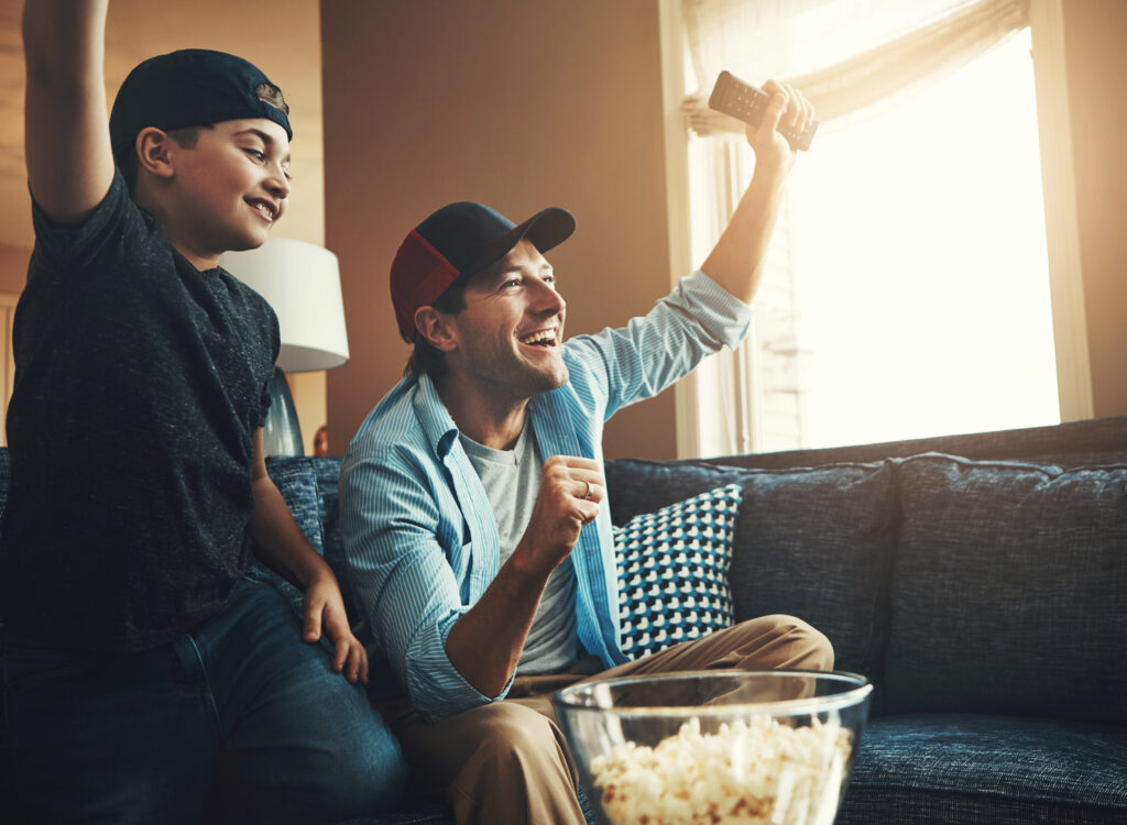 Tv remote, popcorn and man with kid in celebration for network, subscription and bonding at home. Dad, son and happy with fist in success for sports, winning and watching free cable tv live broadcast together.
