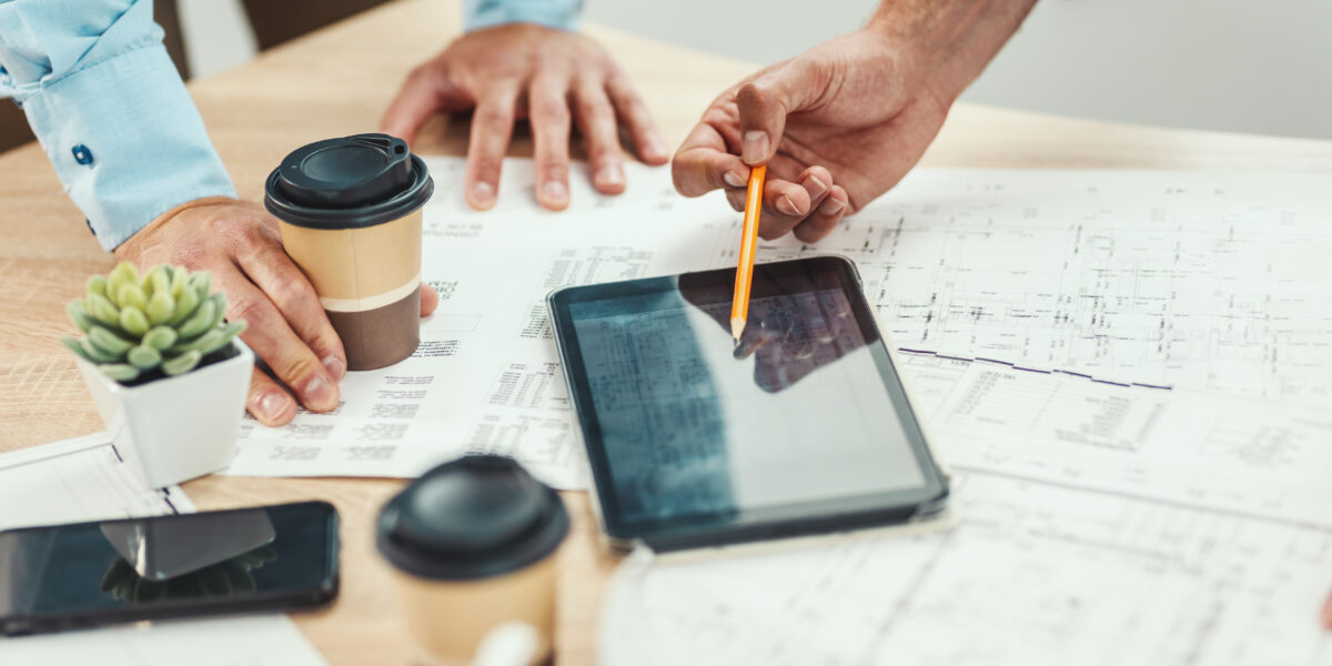 Two professionals leaning over a desk, reviewing tablet plans displayed on a digital tablet while discussing architectural blueprints and documents, with coffee cups nearby.