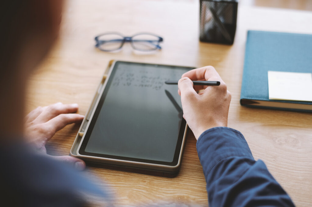 An individual writing tablet plans on a digital tablet using a stylus, focusing on planning and note-taking, with glasses and a notebook in the background.