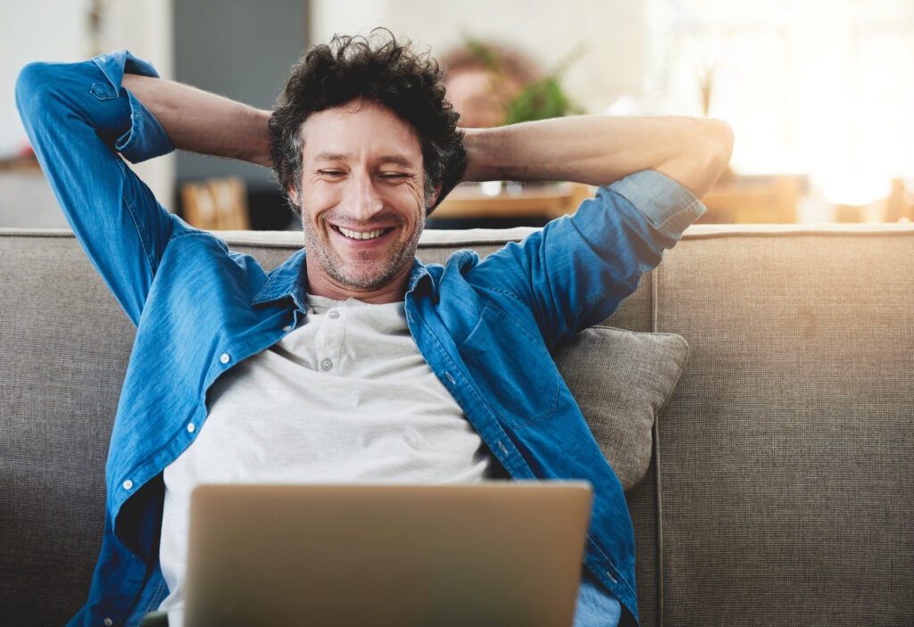 A happy man relaxing on his couch at home, smiling while looking at his laptop screen enjoy his Home Fiber Optic Internet. This image represents the ease and comfort of accessing a fast home fibre optic connection for uninterrupted browsing or work from home.
