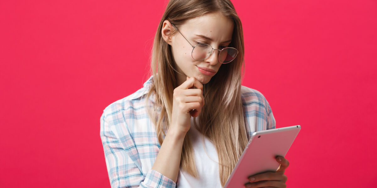 A young woman with glasses, holding a tablet and thinking against a pink background, possibly considering how to "test my ping" for an online task.