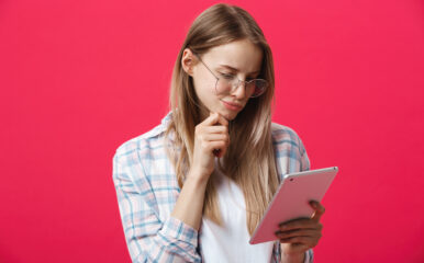 A young woman with glasses, holding a tablet and thinking against a pink background, possibly considering how to "test my ping" for an online task.