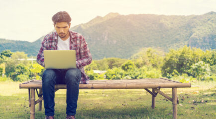 A young man sits on a wooden bench outdoors, using a laptop with a focused expression. He is surrounded by a lush green landscape with mountains in the background, depicting the challenge of getting reliable internet in remote areas. Keywords: how to get internet in rural areas.