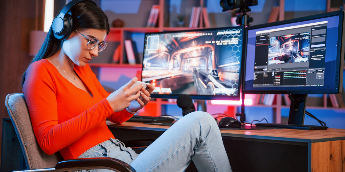 a young woman, likely a game streamer, focused on her smartphone while sitting at a desk with a high-tech setup, including dual monitors displaying a video game stream. This scene could represent the challenge of balancing multiple platforms while addressing a "cancel directv" related to content streaming.
