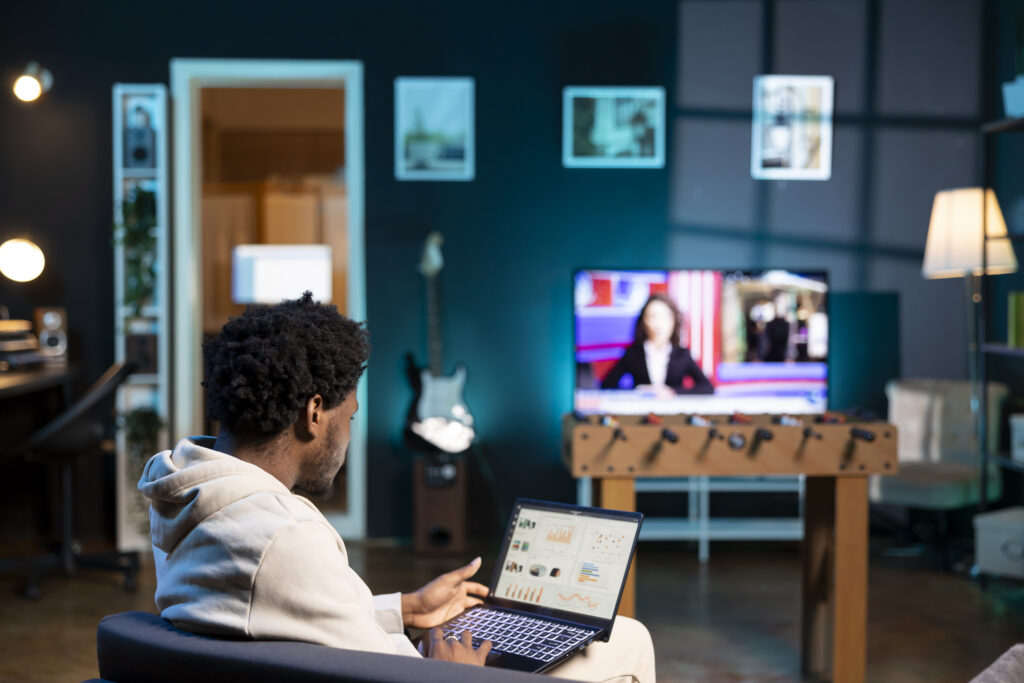 a person sits on a couch reviewing financial data on a laptop screen while a television plays in the background. The individual could be analyzing business performance or responding to a "cancel directv" related to an ongoing project.