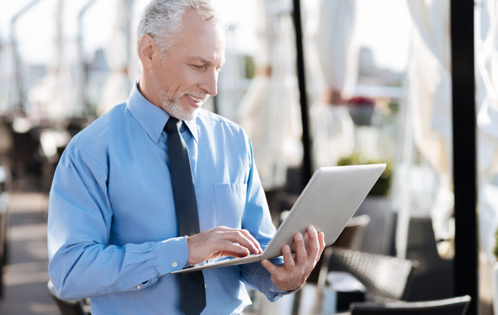 A smiling man in a blue shirt and tie uses his laptop outdoors, highlighting how business fiber optic internet enables seamless, high-speed connectivity for professionals on the go.