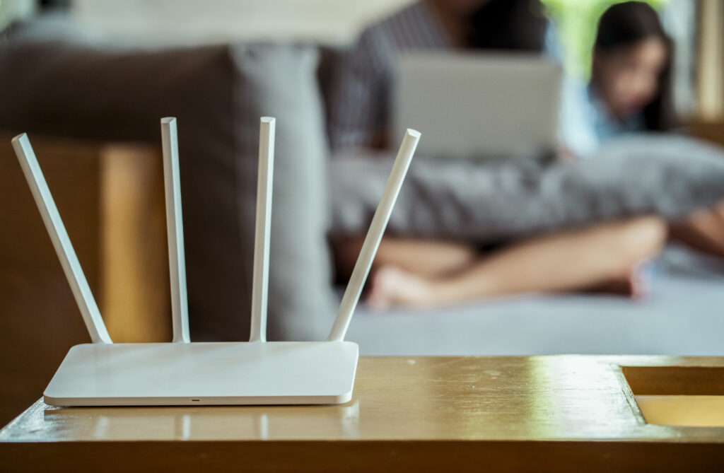 A white WiFi router with four antennas placed on a table in a living room, with a blurred family using a laptop in the background, prompting considerations about "how long do WiFi routers last in shared spaces."
