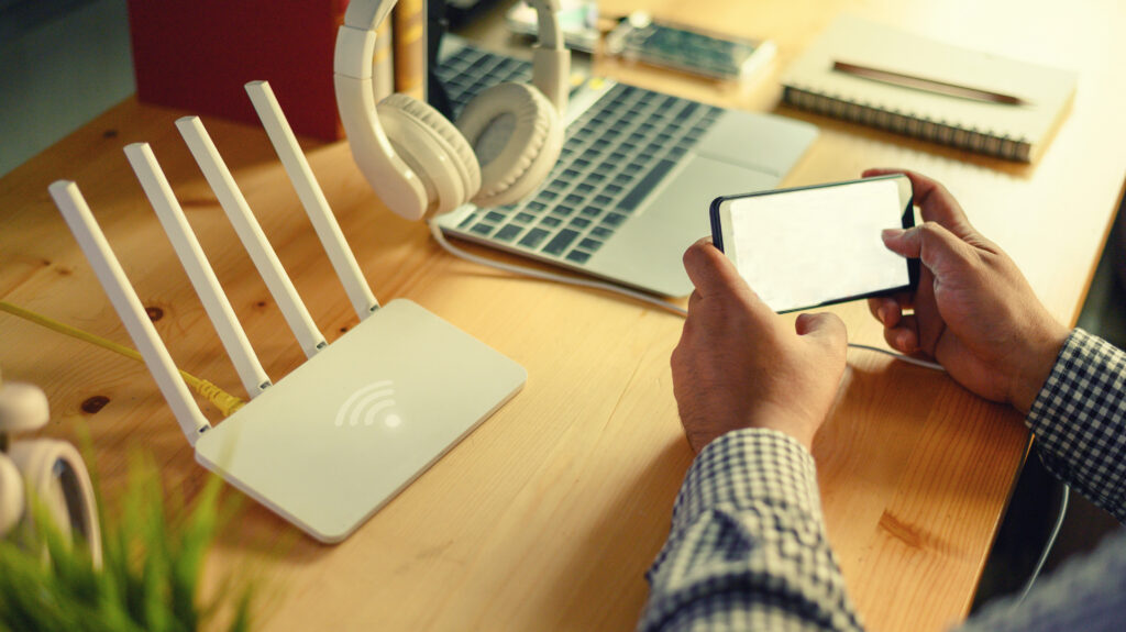  A person holding a smartphone near a WiFi router with four antennas on a wooden desk, surrounded by a laptop and headphones, highlighting the question: "How long do WiFi routers last in home offices?"