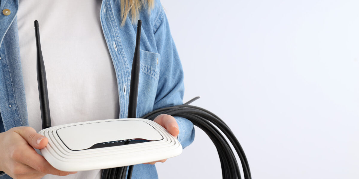 A person holding a white WiFi router with cables against a plain background, representing technical aspects and lifespan inquiries such as "how long do WiFi routers last."