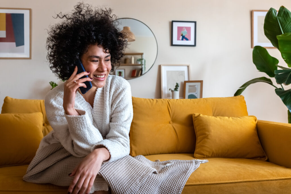  A cheerful woman sitting on a mustard-yellow couch, talking on her smartphone in a cozy living room, illustrating a modern "home phone connect" lifestyle.
