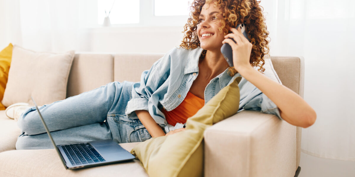 A relaxed woman lounging on a beige sofa, holding her phone while her laptop rests nearby, representing seamless "home phone connect" integration for work and communication.
