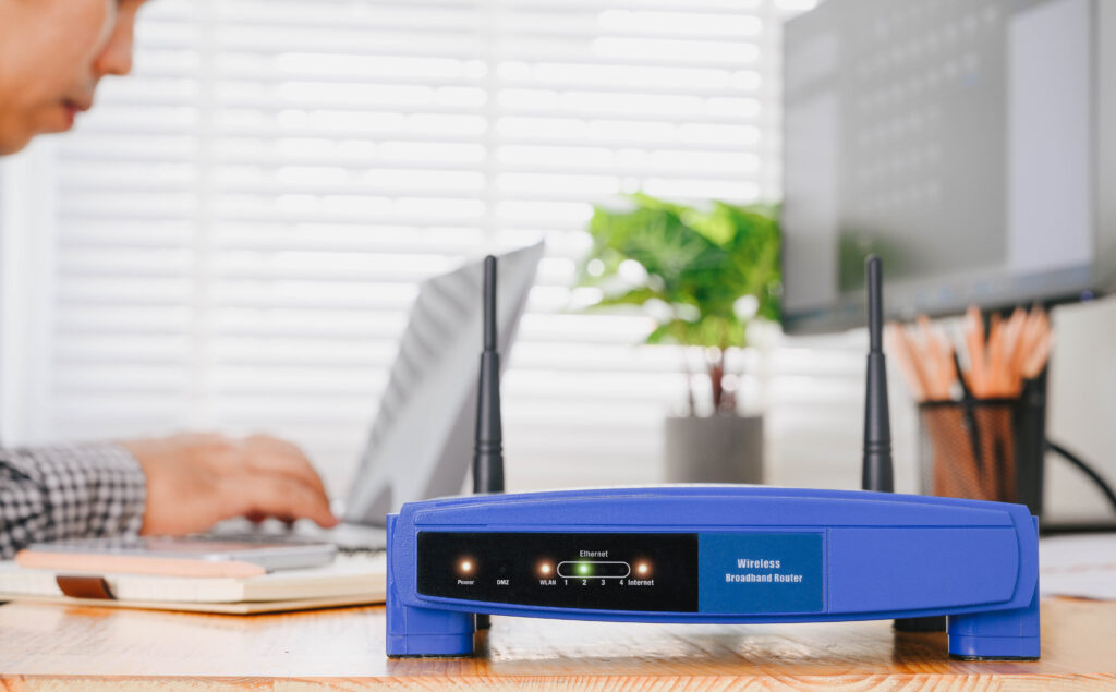A blue wireless broadband router sits on a wooden desk in a bright office space, with a man working on a laptop in the background. "Knowing how often you should replace your router can help you avoid unexpected disruptions in a busy work environment."