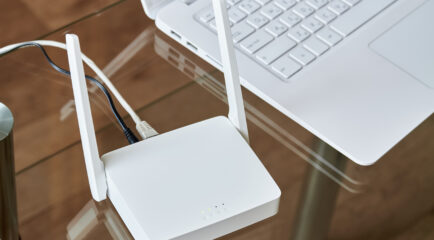 A close-up of a white internet box for home with antennas and ethernet cables connected, sitting next to a laptop on a glass desk for high-speed internet access.