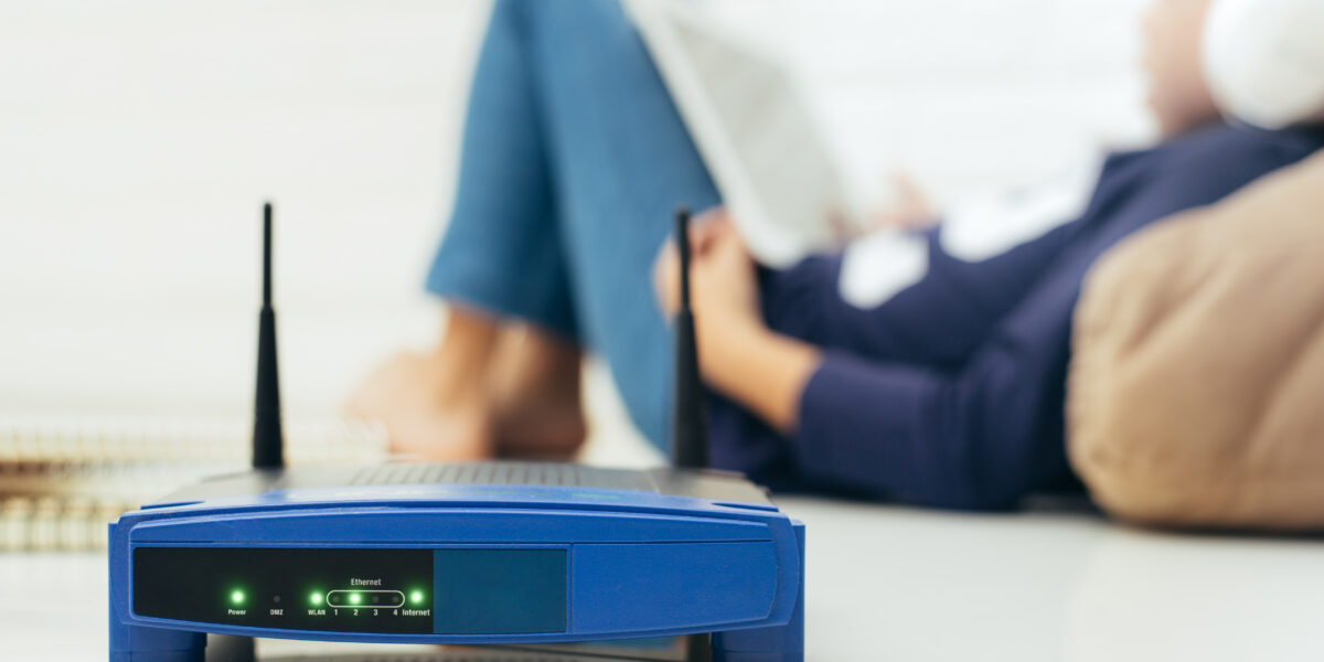 A close-up of a blue wireless router on a white surface, with its indicator lights glowing. In the background, a child is blurred while using a tablet. "Understanding how often you should replace your router is essential for maintaining optimal internet connectivity and performance."